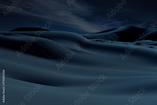 Desert hills and dunes at night under the stars and cloudy sky in blue atmosphere.