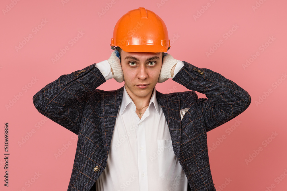 a male engineer in a suit and a protective helmet does not hear anything, covering his ears with his hands on a colored background a male engineer in a suit and a protective helmet does not hear anything, covering his ears with his hands on a colored background