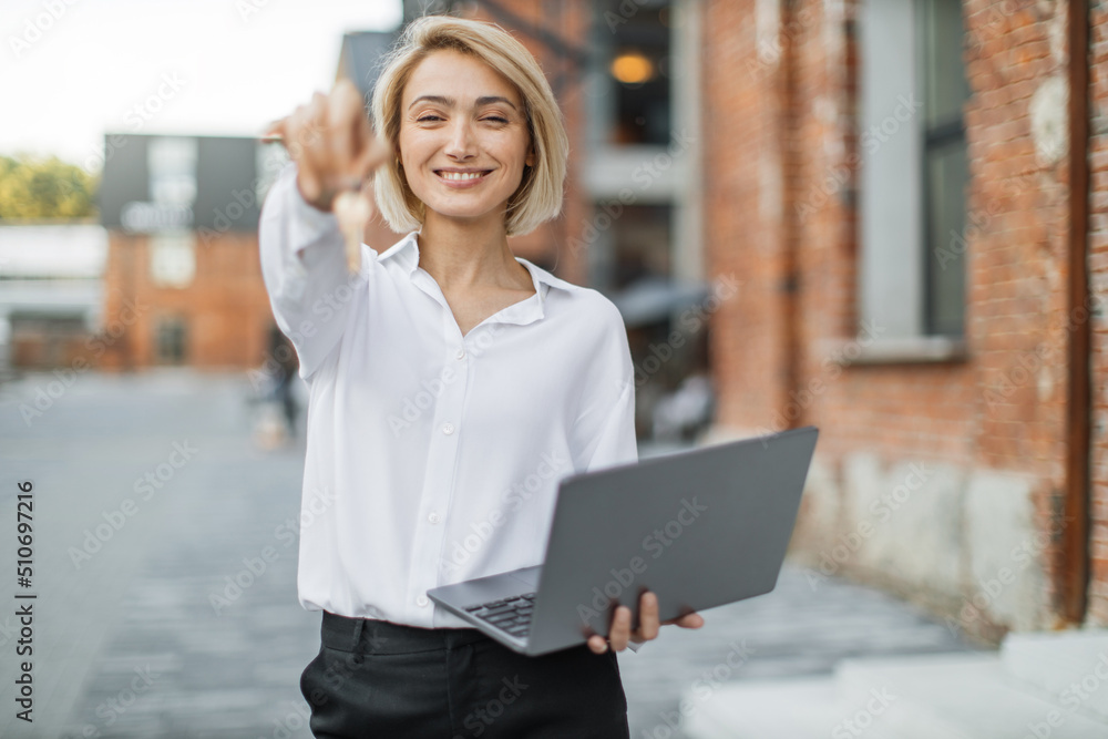 Cheerful young businesswoman in white shirt and black pants holding keys of new flat and laptop, looking at camera and smiling, standing near the building in the middle of the street. Cheerful young businesswoman in white shirt and black pants holding keys of new flat and laptop, looking at camera and smiling, standing near the building in the middle of the street.