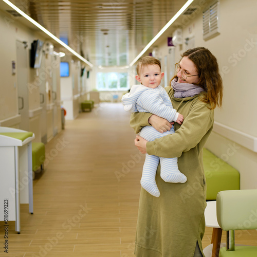 Mother woman with toddler baby boy in her arms visit in the hall clinic. Mom with a child in the corridor of the hospital waiting for a doctor appointment. One year old kid