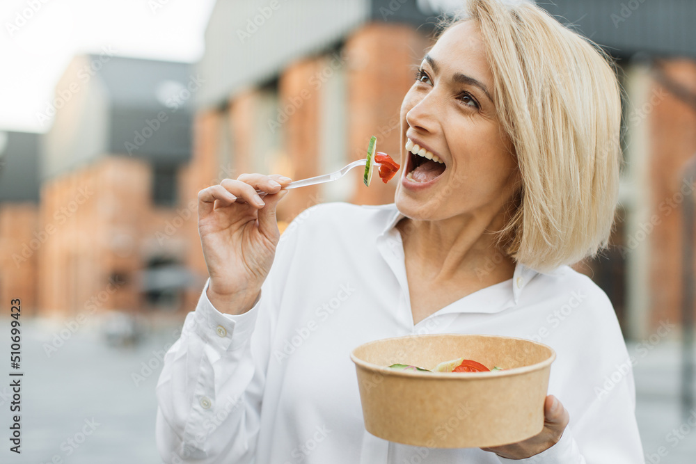 Happy positive business woman eating healthy salad on a break standing over city street background. Female dieting nutrition concept. Attractive smiling girl enjoying veggie meal. Happy positive business woman eating healthy salad on a break standing over city street background. Female dieting nutrition concept. Attractive smiling girl enjoying veggie meal.