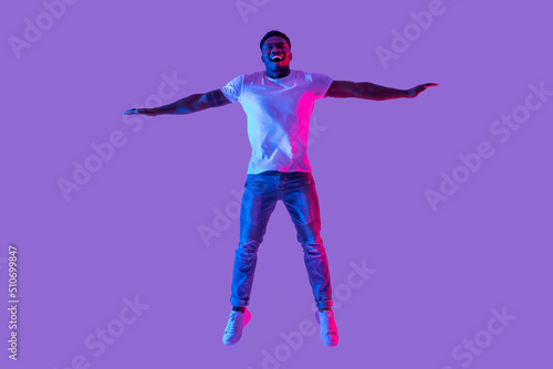 Full length portrait of active energetic young black man jumping in neon light, copy space