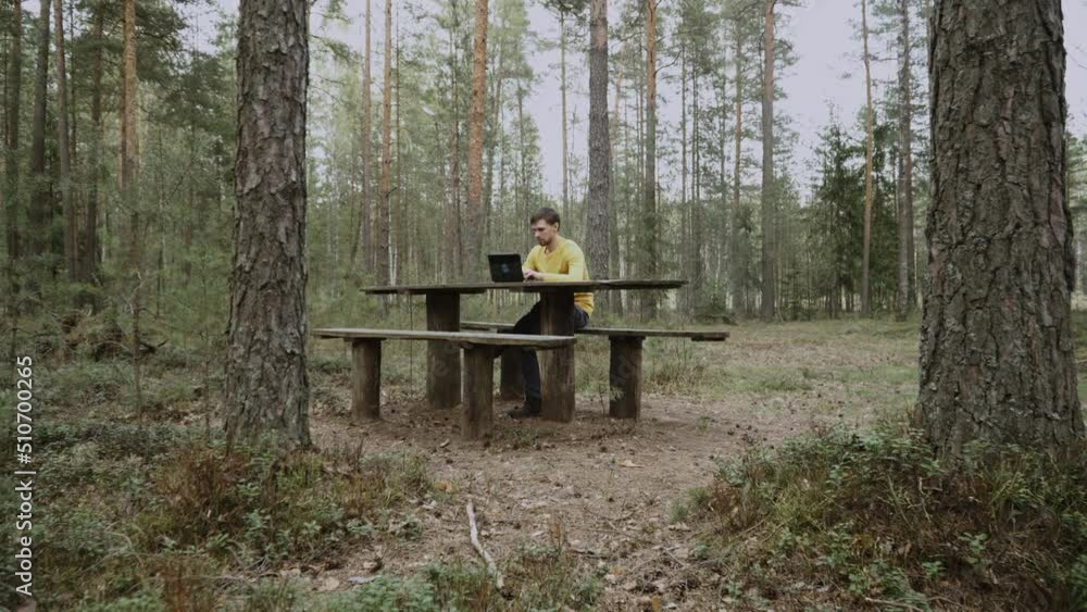 Caucasian man in yellow longsleeve sits at wooden table on bench in green forest and works on laptop. Focused freelancer typing on the keyboard with his hands. Smooth flight camera movement. Caucasian man in yellow longsleeve sits at wooden table on bench in green forest and works on laptop. Focused freelancer typing on the keyboard with his hands. Smooth flight camera movement.