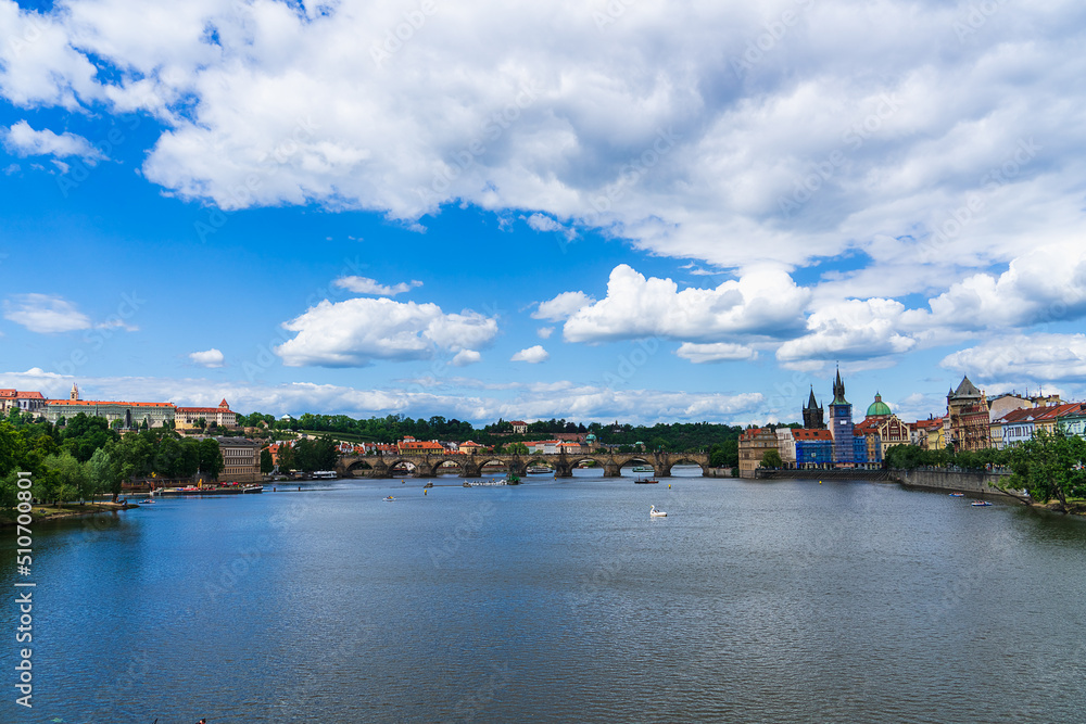 Pleasure boats and pedal boats with vacationers on a sunny day on the Vltava river in Prague, Czech Republic, under a blue sky with white clouds and tall green trees on the shore, european tourism