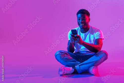 African American guy sitting cross legged with smartphone, surfing social media, chatting to friend in neon light