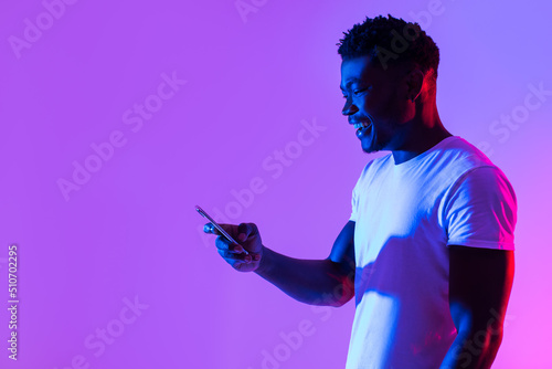 Joyful young black guy using smartphone, studying or working remotely, posting in social media in neon light, copy space