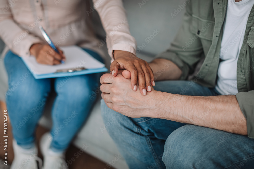 Cropped young black lady psychologist calming european patient and taking notes in clinic