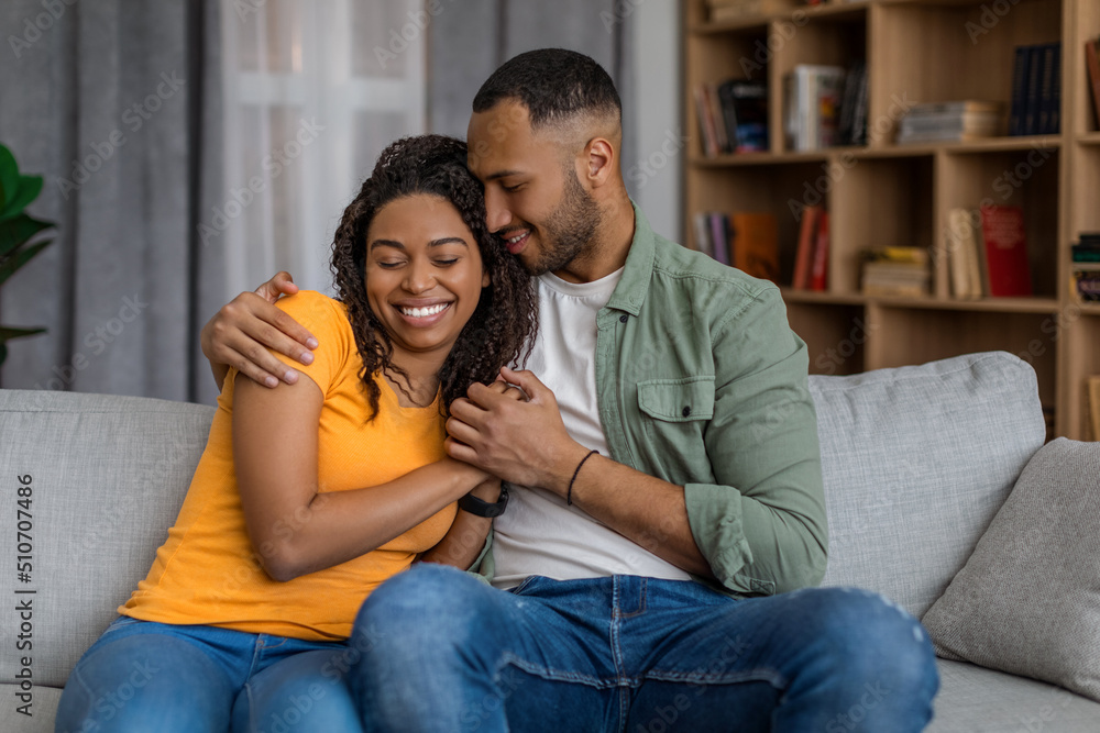 Cute black spouses sitting on sofa, embracing and smiling, enjoying time together at home Cute black spouses sitting on sofa, embracing and smiling, enjoying time together at home