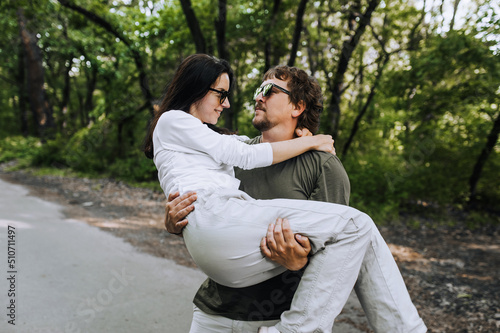 A bearded long-haired stylish man holds his beloved hippie brunette woman in sunglasses in his arms and circles her in nature in a park on the road. Portrait, photo of happy and smiling newlyweds.