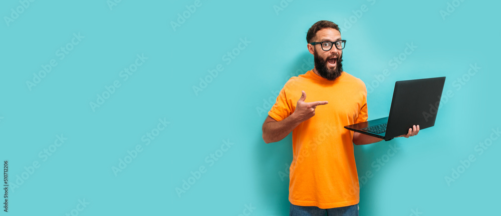 Young crazy bearded charismatic man. Shocked or surprised expression. Laptop concept. Funny promotion poster. Programmer, web developer holding a laptop in his hands and looking at the camera - obrazy, fototapety, plakaty 