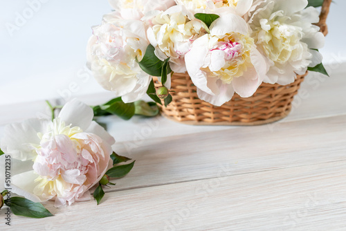 Beautiful fragrant peonies in a wicker basket on a white wooden table. Seasonal flowers as a gift for a birthday, wedding decoration or other celebration © LariBat