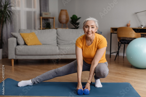 Happy senior woman warming up, doing legs stretching exercises while working out at home on fitness mat