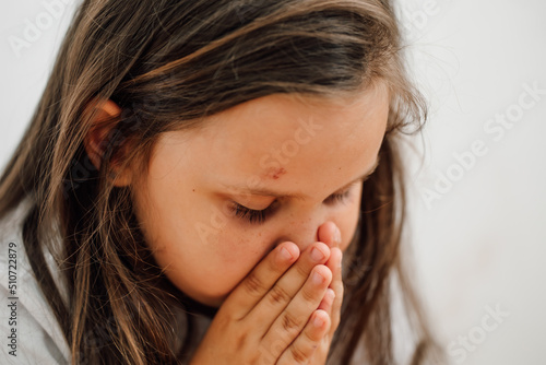  portrait of little sad fearful injured girl with long dark hair sitting with knees up, covering mouth. Protest against domestic violence, abuse, fighting, conflict, bullying. Vertical.