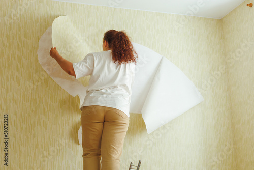 Obraz na plátně Back view of buxom woman with long curly red hair hair wearing white T-shirt, beige trousers, standing on chair, tearing off yellow wallpaper from wall