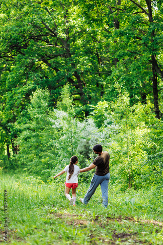 Back view of happy family walking in park forest around green trees, having fun. Little cheerful daughter holding hand of middle-aged bearded man father. Love, summer activities, travelling. Vertical. © Юля Бурмистрова