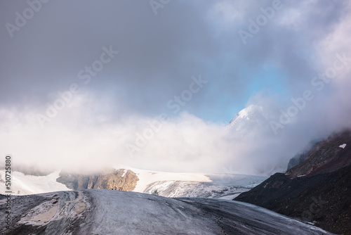 Scenic mountain landscape with large glacier in low clouds in sunrise colors. Colorful view to glacier tongue in morning sunlight in low clouds. Atmospheric high mountain scenery at early morning.