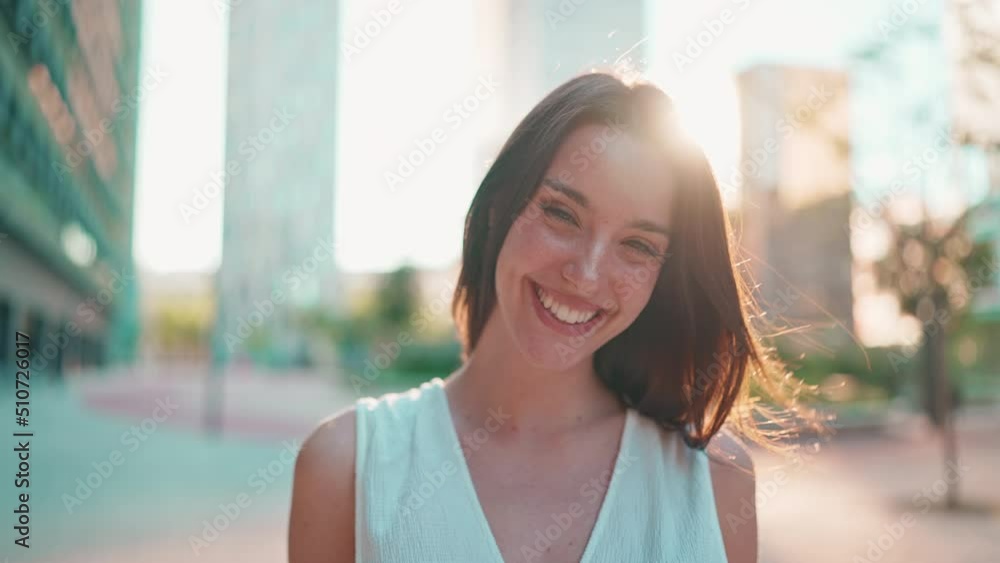 Cute young woman with freckles and dark loose hair wearing white top and jeans is smiling on modern city background. Beautiful girl with long eyelashes smiling in the back light Cute young woman with freckles and dark loose hair wearing white top and jeans is smiling on modern city background. Beautiful girl with long eyelashes smiling in the back light