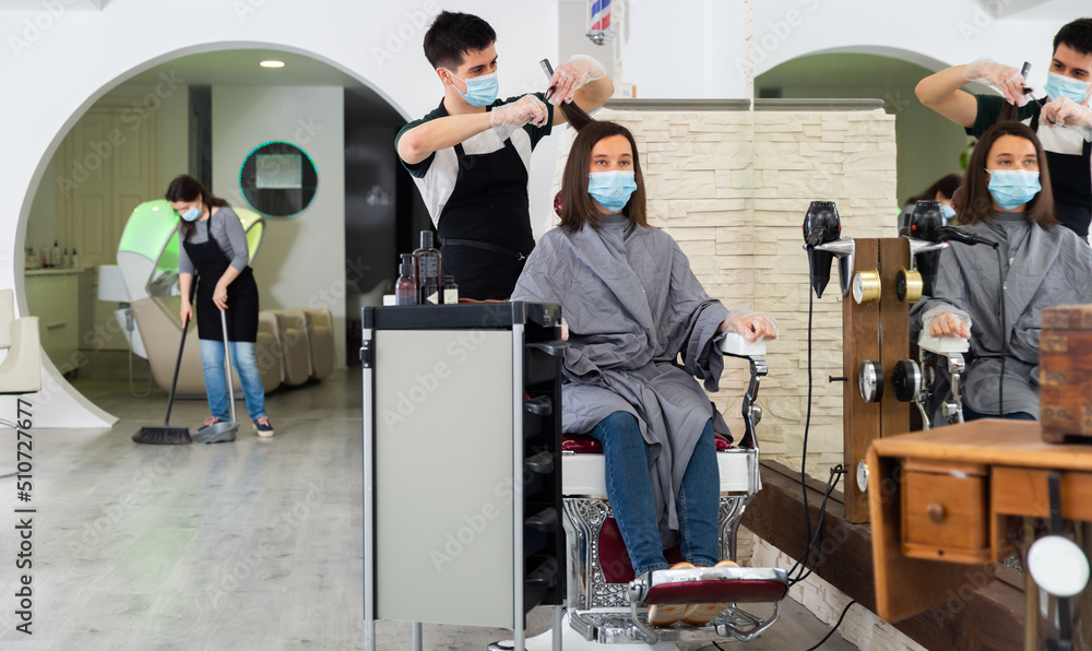 Young man stylist wearing face mask and gloves working in hairdressing salon doing haircut and hair styling for female client