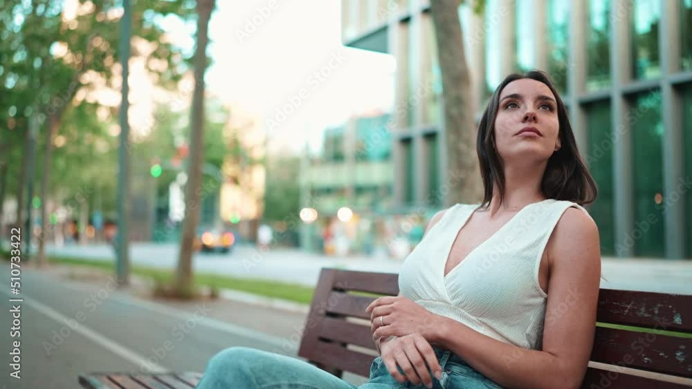 Beautiful woman with freckles and dark loose hair wearing white top sits on bench and looks around. Cute girl is sitting modern city background Beautiful woman with freckles and dark loose hair wearing white top sits on bench and looks around. Cute girl is sitting modern city background
