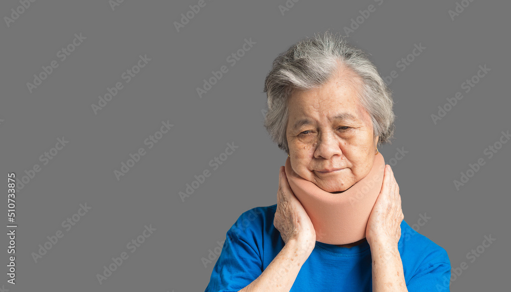 Portrait of a senior woman wearing a soft neck collar because suffering neck pain and neck tilt while standing on a gray background Portrait of a senior woman wearing a soft neck collar because suffering neck pain and neck tilt while standing on a gray background
