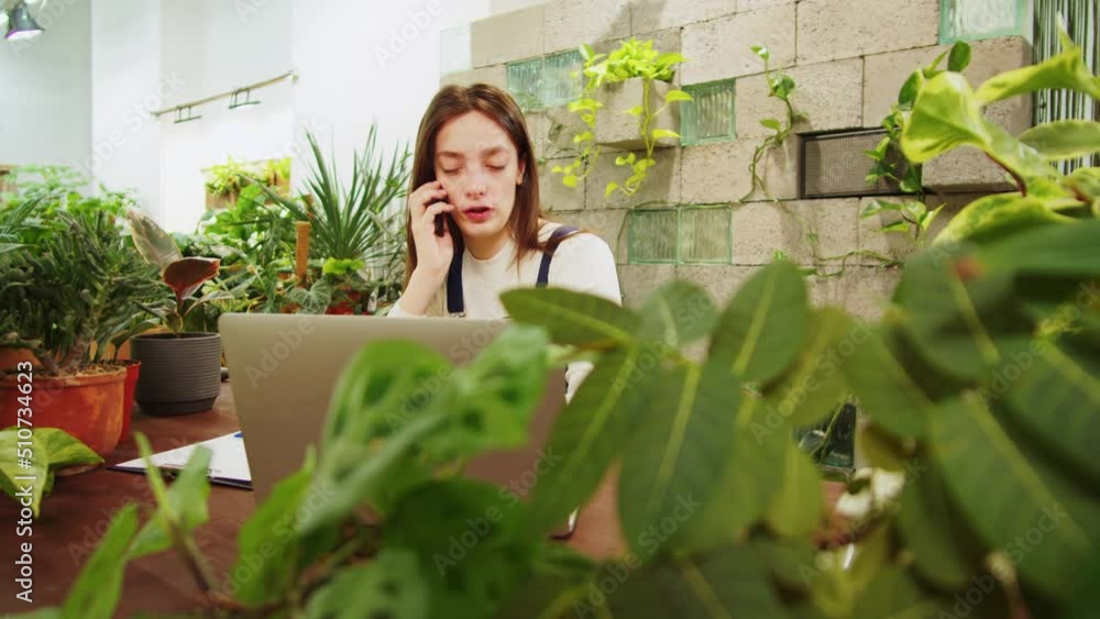 Florist woman calling on phone. Greenhouse manager speaking on smartphone, happy female botanist using laptop close-up, small business owner. Gardener working in flower shop, plant store. Florist woman calling on phone. Greenhouse manager speaking on smartphone, happy female botanist using laptop close-up, small business owner. Gardener working in flower shop, plant store.