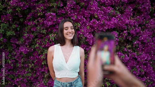 Beautiful woman with freckles and dark loose hair wearing white top poses wall in bloom with purple flowers. Pretty girl is photographed on smartphone