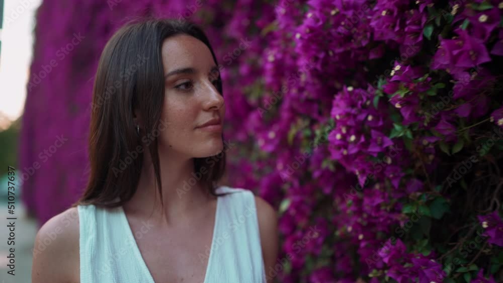 Beautiful woman with freckles and dark loose hair wearing white top walks along wall blooming with purple flowers. Cute girl sniffing purple flowers Beautiful woman with freckles and dark loose hair wearing white top walks along wall blooming with purple flowers. Cute girl sniffing purple flowers