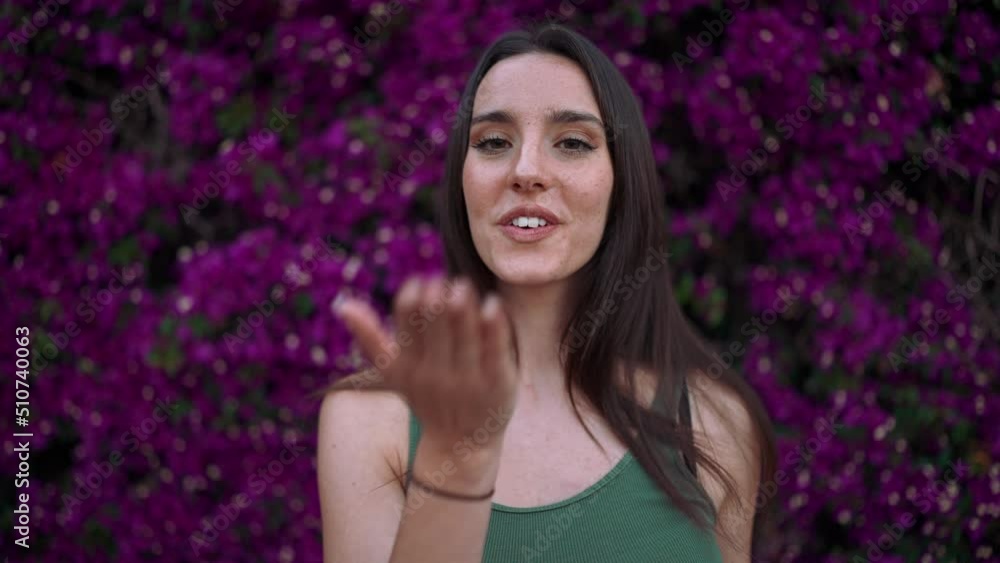 Beautiful woman with freckles and dark loose hair wearing green top poses wall in bloom with purple flowers. Pretty girl sends an air kiss and smiles while looking at the camera Beautiful woman with freckles and dark loose hair wearing green top poses wall in bloom with purple flowers. Pretty girl sends an air kiss and smiles while looking at the camera