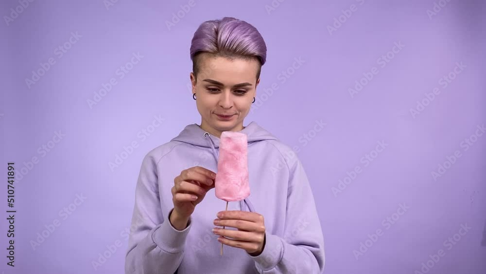 Cheerful young adult woman holding cotton candy on stick, looking at sweet good in anticipation, licking lips, getting piece of candy floss, eating with pleasure isolated on purple background indoors Cheerful young adult woman holding cotton candy on stick, looking at sweet good in anticipation, licking lips, getting piece of candy floss, eating with pleasure isolated on purple background indoors