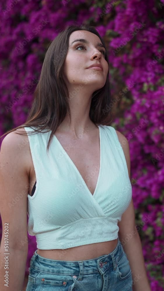 VERTICAL VIDEO: Beautiful woman with freckles and dark loose hair wearing white top walks along wall blooming with purple flowers. Cute girl looking at purple flowers VERTICAL VIDEO: Beautiful woman with freckles and dark loose hair wearing white top walks along wall blooming with purple flowers. Cute girl looking at purple flowers