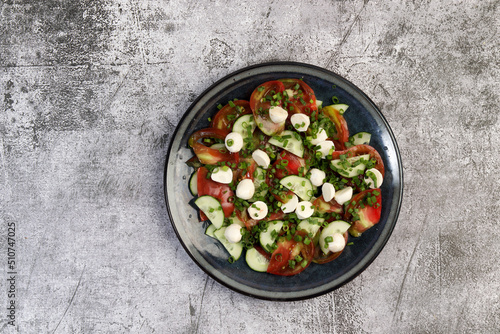 Summer homemade Tomato, mozzarella and cucumbers salad on a round plate on a dark gray background. Top view, flat lay