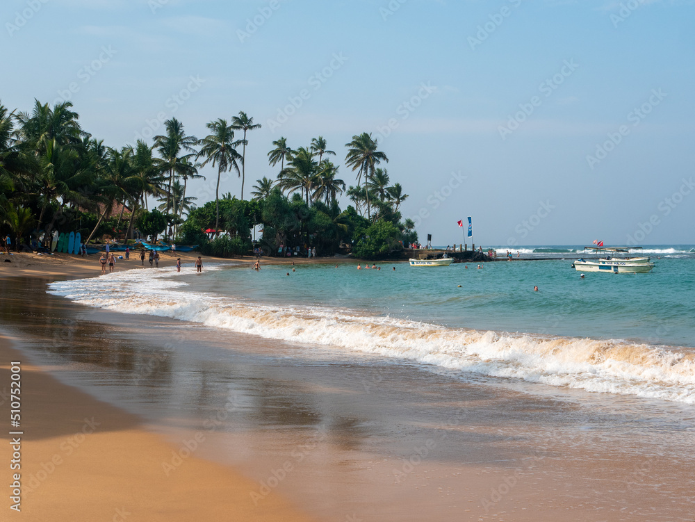 Hikkaduwa, Sri Lanka - March 4, 2022: Beautiful view of the beach in Hikkaduwa with bright green palm trees. People relax and swim in the Indian Ocean