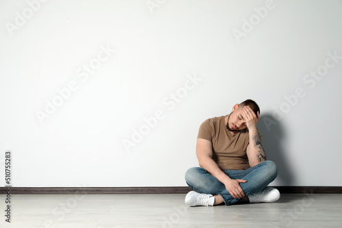 Sad young man sitting on floor near white wall indoors, space for text © New Africa