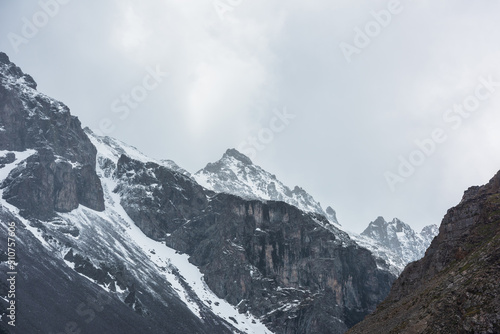 Awesome landscape with high snowy mountain peaked top with sharp rocks in cloudy sky. Dramatic view to snow mountain pointed peak in rainy weather. Atmospheric scenery with white snow on black rocks.