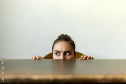 Funny indoor picture of frowning student girl with hair knot and guarded look hiding behind table, concentrated eyes looking aside, listening attentively to strange sounds and noise photo