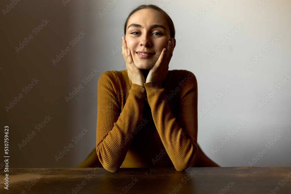 Funny charming dreamy female with lovely charismatic smile sitting at table in turtleneck, resting head on hands, isolated on background of office or classroom, young teacher having rest after work Funny charming dreamy female with lovely charismatic smile sitting at table in turtleneck, resting head on hands, isolated on background of office or classroom, young teacher having rest after work