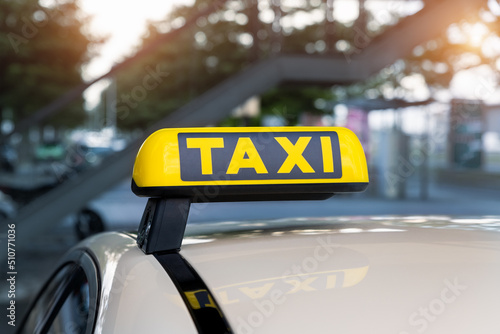Close-up detail yellow taxi symbol on cars roof stand waiting at parking of airport terminal or railway station against park warm evening bokeh sunlight. Urban street transportation comfort service