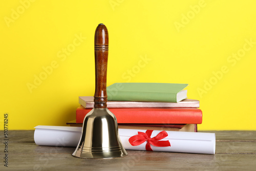 Golden school bell, diploma and books on wooden table against yellow background
