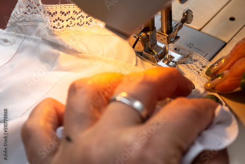Fototapeta Naklejka Na Ścianę i Meble -  Close up view of sewing process. Female hands stitching white fabric on professional manufacturing machine at workplace. Seamstress hands holding textile for dress production. Light blurred background
