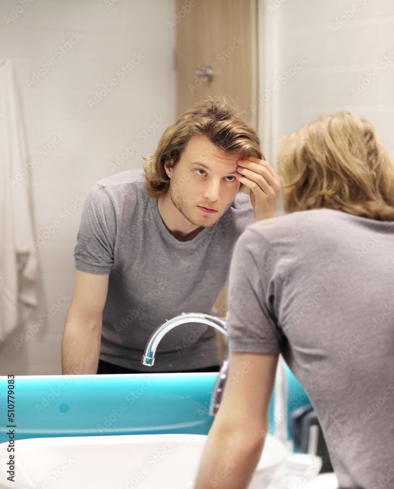 Obraz premium young man looking in the mirror,combing his hair,looking at problems on face.