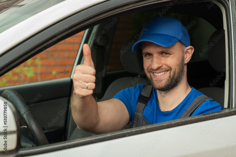 Man in uniform sits on driver seat looking at camera and showing thumb-up closeup. Repairman smiles satisfied with result of repair after fixing car Man in uniform sits on driver seat looking at camera and showing thumb-up closeup. Repairman smiles satisfied with result of repair after fixing car