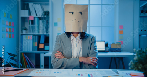 Young Asian businesswoman sit on desk wear draw sad mask paper craft bag feel bad mood with fail work project in office at night Fototapet