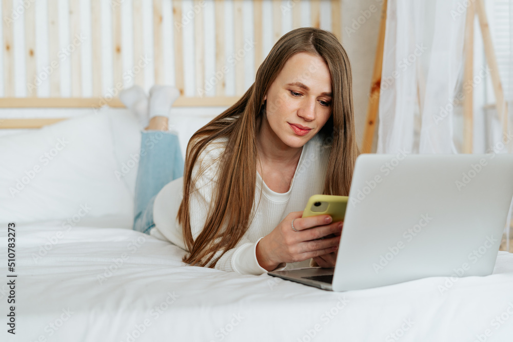 Freelance woman texting over cellphone and having a video call chat online shopping, lying on the white bed. Happy relaxed girl woking from home office. Distance learning online education and work. Freelance woman texting over cellphone and having a video call chat online shopping, lying on the white bed. Happy relaxed girl woking from home office. Distance learning online education and work.