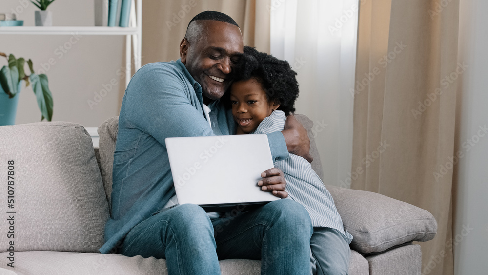 Adult african american male freelancer sitting on sofa working on laptop little daughter interferes with dad work asks for attention loving father hugging kid girl parenthood parental responsibilities Adult african american male freelancer sitting on sofa working on laptop little daughter interferes with dad work asks for attention loving father hugging kid girl parenthood parental responsibilities