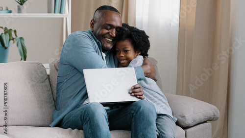 Adult african american male freelancer sitting on sofa working on laptop little daughter interferes with dad work asks for attention loving father hugging kid girl parenthood parental responsibilities