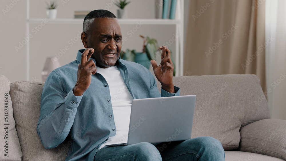 Mature african american man sitting at home looking at laptop screen with hope crossed fingers praying for good luck hopeful wishes victory success asking fortune asks for win believing in winning Mature african american man sitting at home looking at laptop screen with hope crossed fingers praying for good luck hopeful wishes victory success asking fortune asks for win believing in winning