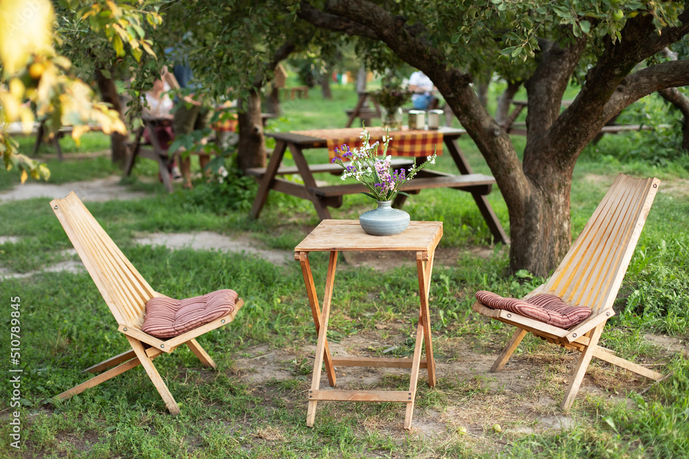 Wooden chairs and table table in summer orchard. Wooden outdoor furniture set for Picnic in garden. Cozy Interior Courtyard with table and sun loungers. Garden furniture for leisure time in nature.	