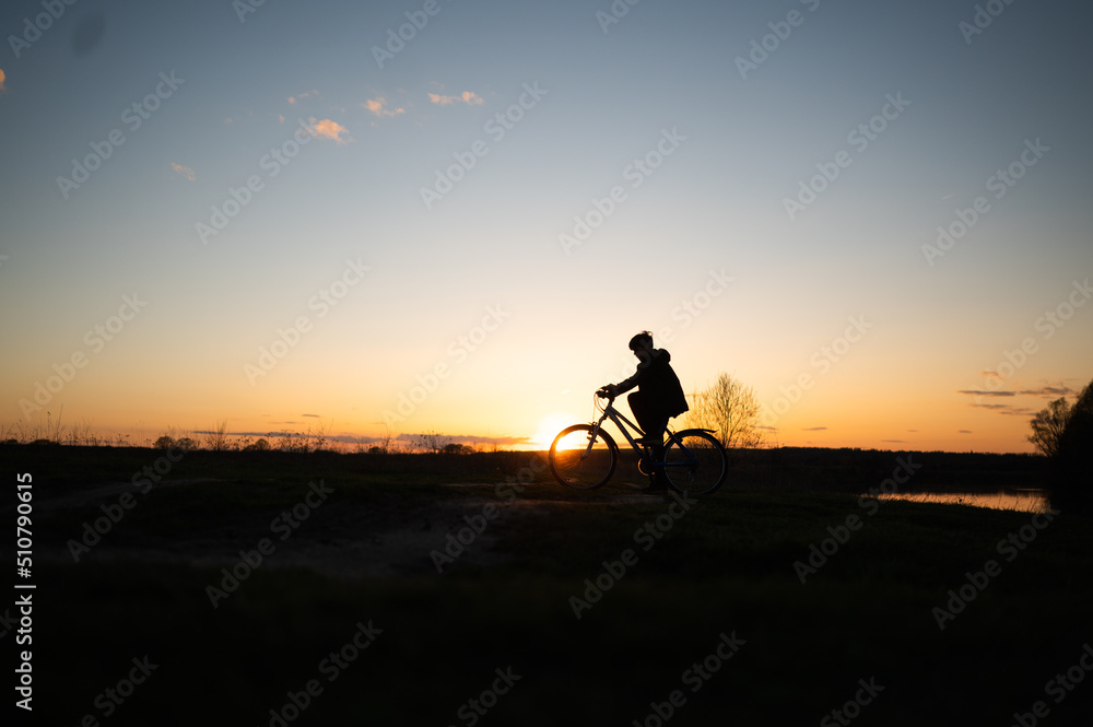 The silhouette of a boy on a bicycle riding along the river. Silhouette of a cyclist on a hill beautiful colorful sky and clouds in the background. Silhouette against the sunset