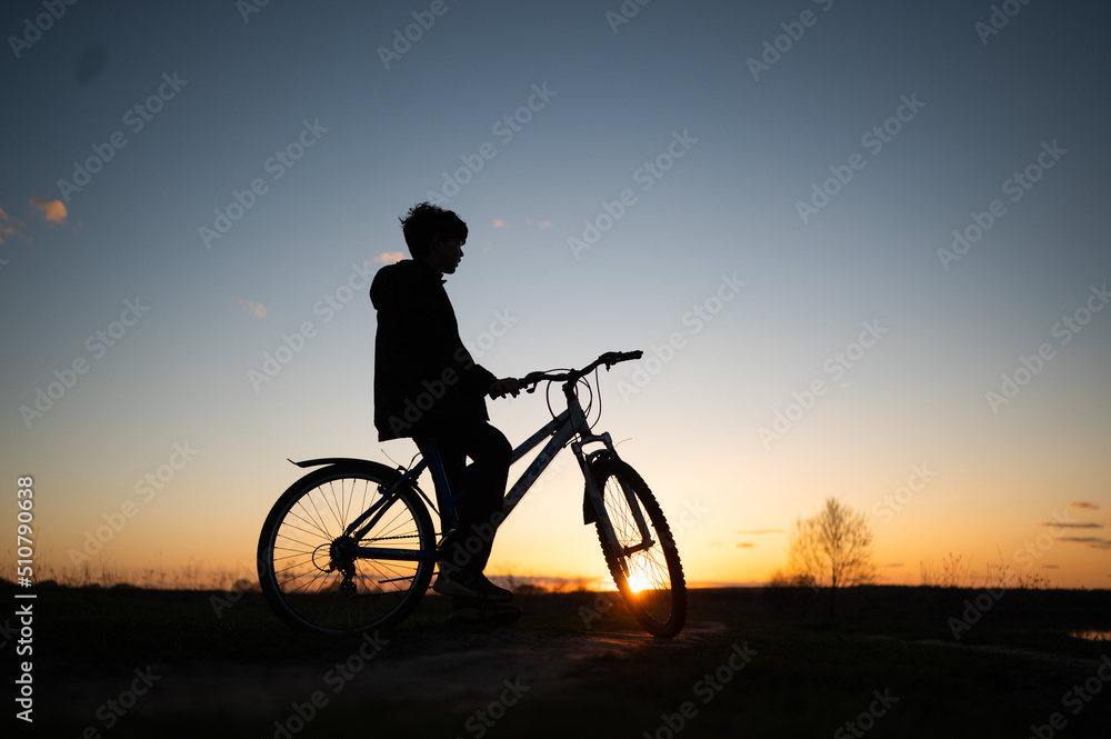 Obraz premium The silhouette of a boy on a bicycle riding along the river. Silhouette of a cyclist on a hill beautiful colorful sky and clouds in the background. Silhouette against the sunset