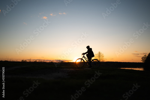 The silhouette of a boy on a bicycle riding along the river. Silhouette of a cyclist on a hill beautiful colorful sky and clouds in the background. Silhouette against the sunset